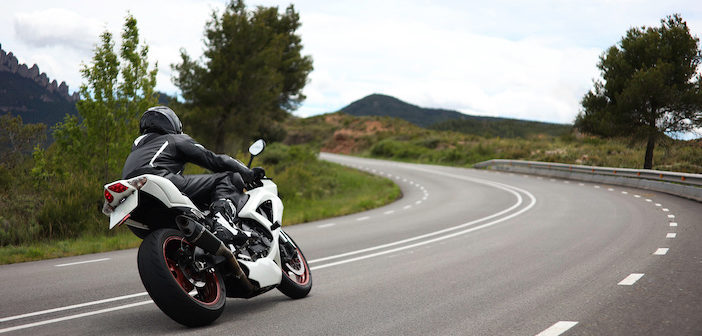 Biker in a black uniform driving a white sports motorcycle on a curvy road in the middle of a green meadow with trees and with mountains in the background on a cloudy day